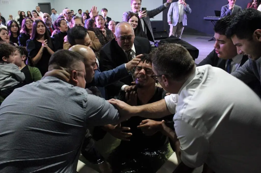 Hombre recibiendo el bautismo en la iglesia Firstborn Ministerios en Rockford, IL, rodeado de ministros y fieles en un momento de fe y transformación espiritual."