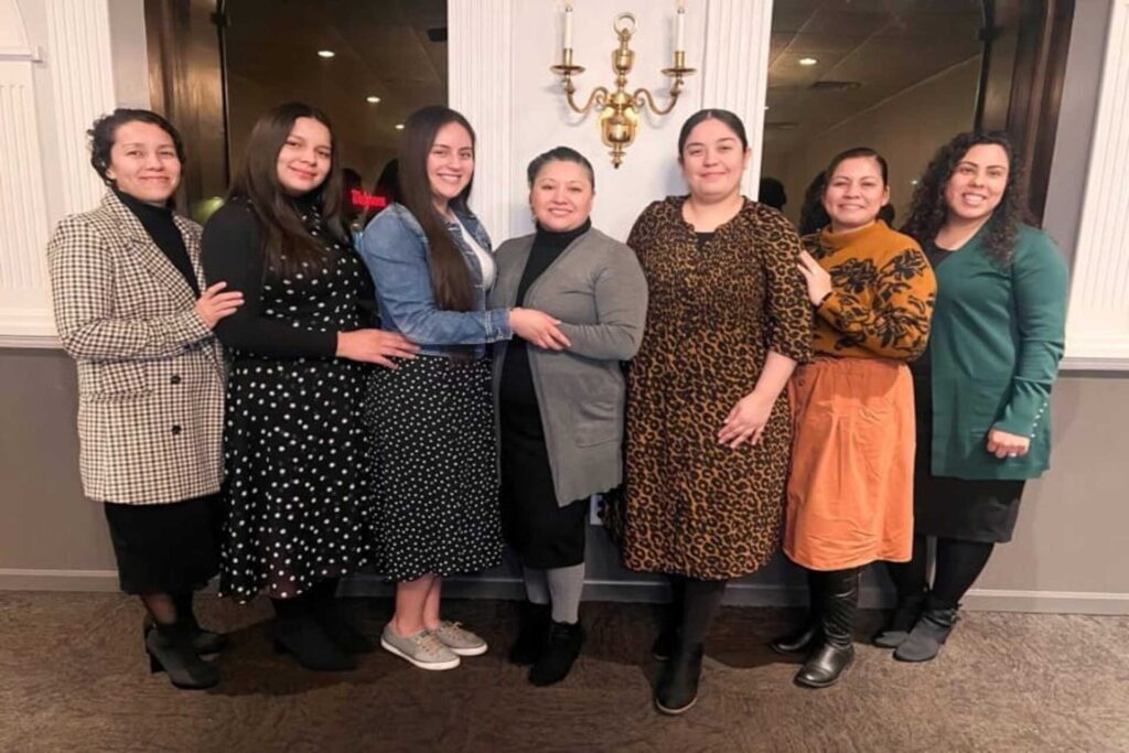 A group of seven women from Firstborn Ministerios posing together at a church event, dressed in elegant and modest attire, smiling in a warmly lit indoor setting.